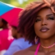 A person with curly hair and hoop earrings looks toward the camera, surrounded by others wearing pink and blue attire in an outdoor setting.