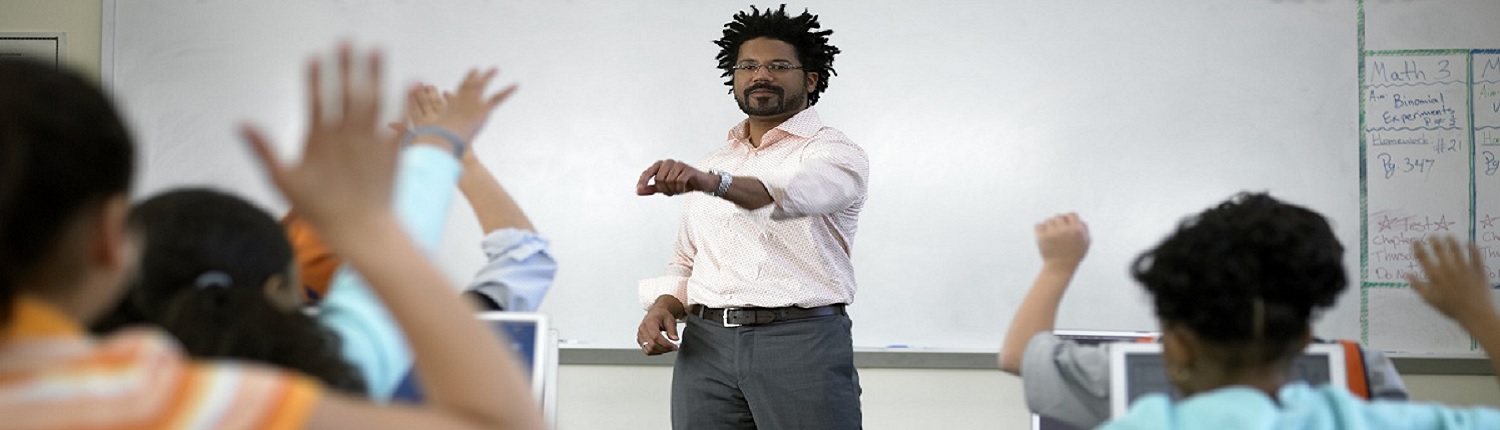 A teacher stands at the front of a classroom with a whiteboard, as students raise their hands to ask questions.