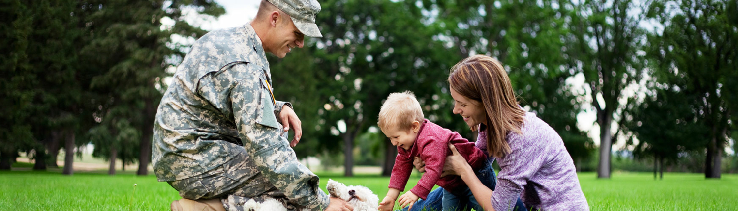 A soldier in uniform kneels on grass, smiling at a young child and a woman, as they all pet a dog in a park setting.