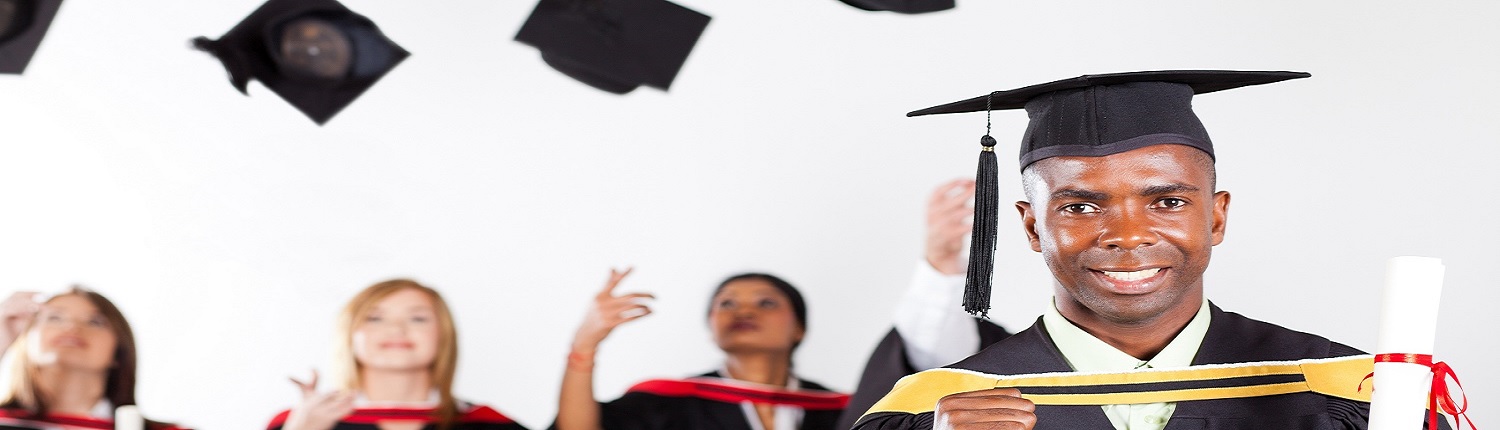 A graduate in a cap and gown smiles while holding a diploma. In the background, other graduates toss their caps in the air.