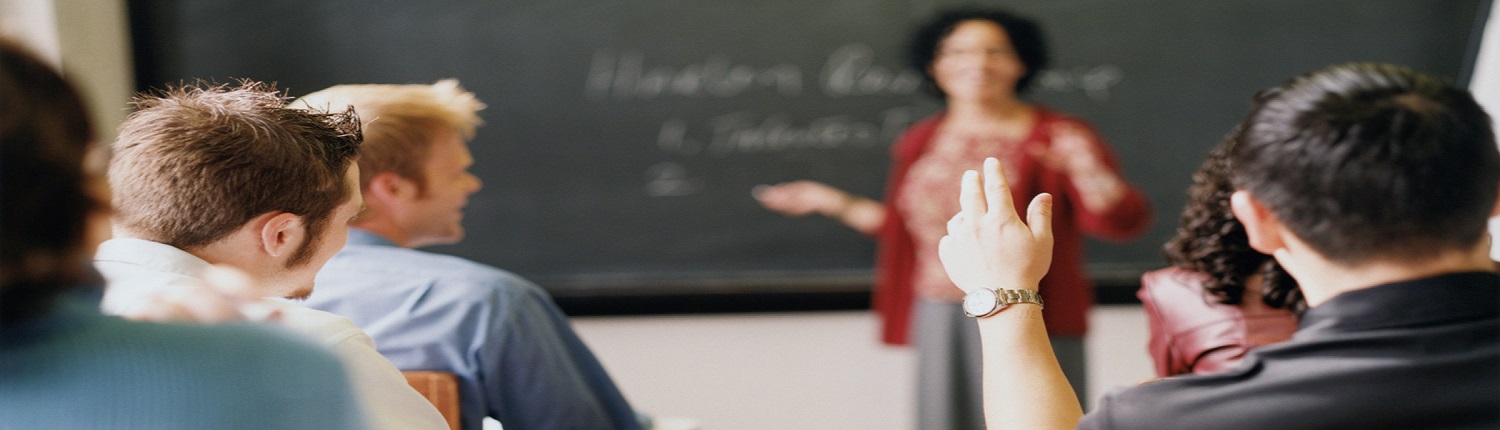 Students in a classroom facing a teacher at the blackboard, one student raising a hand.